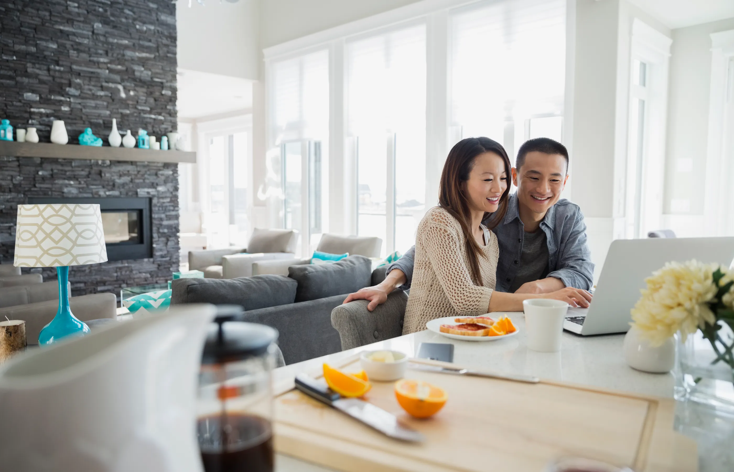 A couple is sitting at their kitchen counter, eating food, while reviewing information on their laptop. 